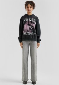 Czarna bluza z kapturem z graficznym nadrukiem przedstawiającym rękę i tekst, w połączeniu z szarymi dżinsami o wysokim stanie; gładki materiał, luźny krój i długie rękawy.