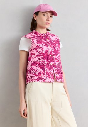 Γυναίκα που φοράει ροζ φλοράλ γιλέκο με διπλή ραφή πάνω από λευκό t-shirt, εκρού παντελόνι με φαρδιά μπατζάκια και ροζ καπέλο μπέιζμπολ, στέκεται μπροστά σε ανοιχτό γκρι τοίχο.