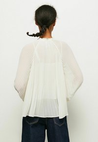 Vit plisserad blus med långa, flöjtande ärmar och hög halsringning, kombinerad med mörkblå jeans. Lätt, genomskinligt tyg.