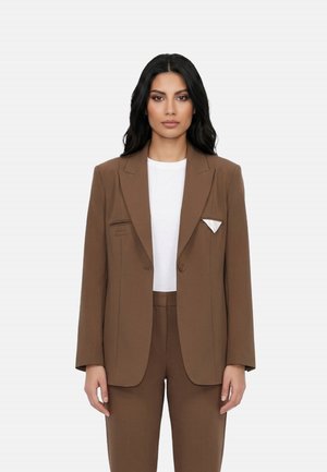 Donna con capelli lunghi e neri che indossa un blazer marrone su misura e pantaloni sopra una camicia bianca, in piedi davanti a uno sfondo chiaro e uniforme.