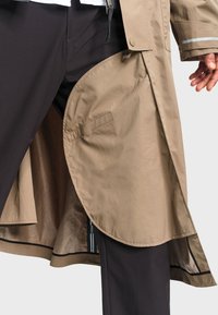 Trench-coat beige partiellement ouvert montrant la doublure intérieure, porté avec un pantalon noir, une main détendue le long du corps sur un fond clair.