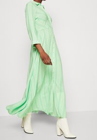 Robe maxi boutonnée verte à motif chevrons, avec manches longues et silhouette fluide, associée à des bottes blanches à talons hauts.