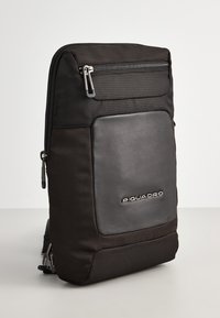 Zaino di stoffa nera con superficie texturizzata, dotato di una tasca frontale in pelle liscia e chiusura superiore con zip. Include logo marchiato.
