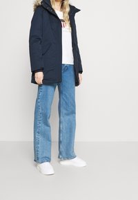 Tmavě modrá parka bunda s kapucí lemovanou umělým kožíškem, zapínáním na zip a bočními kapsami, nošená přes bílý svetr a doplněná světle modrými džínami a bílými teniskami.