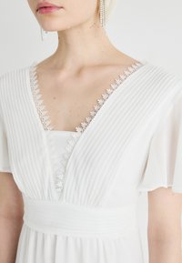 Valge kleit, millel on volditud kangas, pitsaktsentid kaeluses ja sobiv vöökoht. Pehmed, voolavad varrukad lisavad elegantsi.