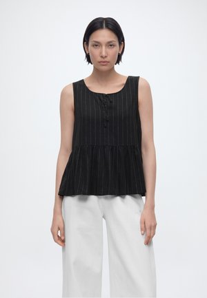 Kvinne med kort mørkt hår iført en ermeløs svart bluse med nålestriper og løse hvite bukser, stående mot en ensfarget bakgrunn.