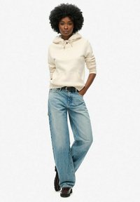 Sudadera beige con capucha, bolsillo frontal y cordones, combinada con jeans de mezclilla azules sueltos. El modelo lleva un cinturón negro y botas marrones hasta el tobillo.