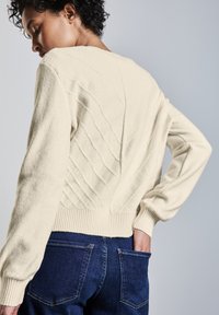 Strickpullover in Creme mit strukturiertem Wellenmuster auf dem Rücken, geripptem Saum und langen Ärmeln. Getragen mit blauen Jeanshosen mit Taschen.
