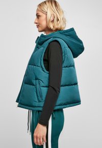Gilet en duvet turquoise avec capuche, devant zippé et poches latérales. Présente une texture lisse et brillante ainsi qu'un design court et carré.