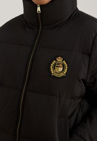 Μαύρο μπουφάν puffer με ψηλό γιακά, διακοσμημένο με χρυσό κεντημένο έμβλημα και φερμουάρ κλεισίματος. Ομαλό ύφασμα με σχεδιασμό ραμμένων πάνελ.