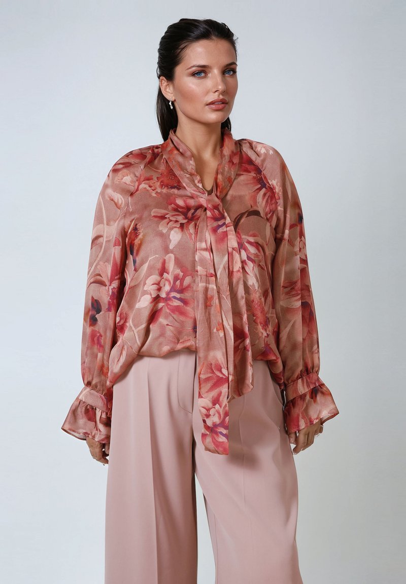 Luftige, transparente Bluse mit floralem Druck in Rosa- und Beigetönen; mit einem gebundenen Kragen und langen Rüschensäumen. Kombiniert mit sanften rosa Hosen.