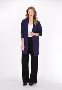 Cardigan lungo viola realizzato in morbido tessuto, con apertura frontale, indossato su una maglietta bianca e pantaloni a gamba larga neri, con scarpe con tacco.