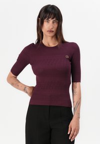 Bordeaux ribbed gebreid top met korte mouwen en een ronde halslijn. Voorzien van een klein gouden embleem op de linkerkant van de borst.