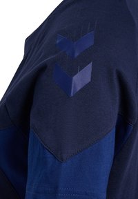 Tummansininen lyhythihainen paita, jossa on teksturoitu kangas. Sisältää tummemmasta sävystä olevan chevron-kuvion olkapäässä ja kontrastivärisen sinisen paneelin.