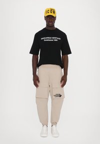 T-shirt noir à manches courtes avec du texte blanc, pantalon cargo beige avec poches, et sneakers blanches, le tout surmonté d'une casquette de baseball jaune avec l'étiquette "ICON."