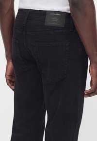 Άνδρας που φοράει μαύρα τζιν Jack & Jones, δείχνει τις πίσω τσέπες και την ετικέτα της μάρκας στη μέση.