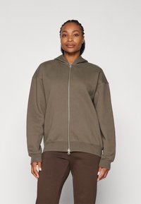 Khaki mikina na zips s kapucňou, dlhými rukávmi a rebrovaným spodkom. Mäkká tkanina s voľným strihom, v kombinácii s hnedými nohavicami.