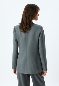 Grijze op maat gemaakte blazer met een gestructureerde pasvorm, voorzien van een strak ontwerp, rechte rugnaad en een gladde textuur. Draagt als onderdeel van een pak.