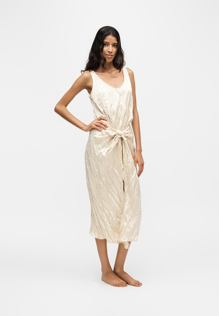 Femme aux longs cheveux foncés porte une robe crème sans manches, longueur genou, avec un nœud à l'avant, debout pieds nus sur un fond blanc.
