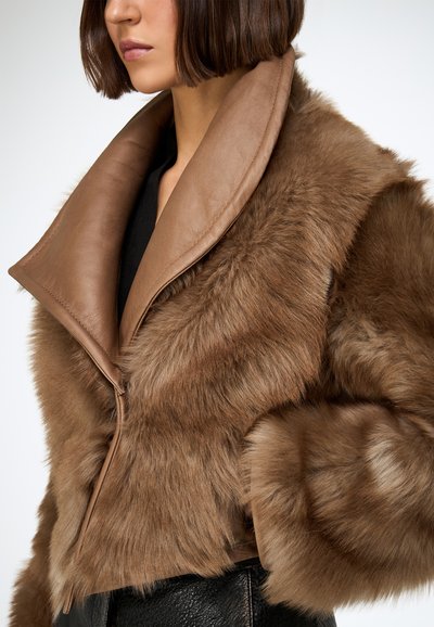 Veste en fourrure marron avec un col large en cuir lisse. Présente une texture moelleuse et une coupe courte, portée avec un vêtement noir élégant en dessous.
