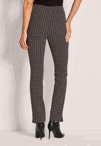 Vrouw in zwart-wit geometrisch patroon slim fit broek en zwarte hoge hakken enkellaarsjes, staand tegen een effen achtergrond.