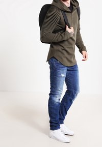 Olivgrön zip-hoodie med ribbade ärmar, som parats ihop med slitna blå jeans och vita sneakers. Svart ryggsäck hängande på vänster axel.