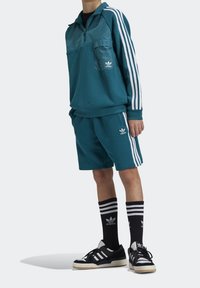 Tyrkysová mikina se zipem do poloviny, náprsní kapsou a bílými pruhy, zkombinovaná s odpovídajícími šortkami. Černobílé pruhované ponožky a tenisky.