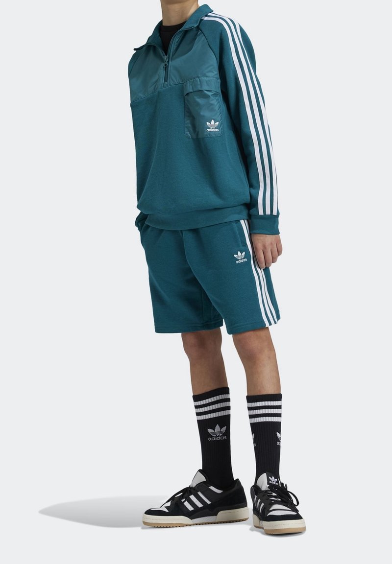 Tyrkysová mikina se zipem do poloviny, náprsní kapsou a bílými pruhy, zkombinovaná s odpovídajícími šortkami. Černobílé pruhované ponožky a tenisky.