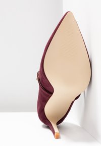 Chaussure à talon haut en daim bordeaux à bout pointu reposant sur le côté, montrant la semelle lisse beige et l'extrémité fine du talon.