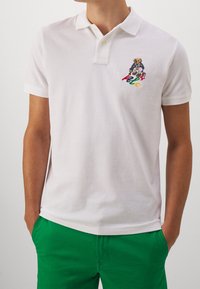 Bílá polokošile z texturované bavlny s dvouknoflíkovým límečkem a barevným motivem medvěda a vyšitým nápisem "Polo" na levé straně hrudi.