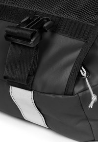 Svart tygväska med en texturerad rem, plastspänne och en vit horisontell rand. Detaljer inkluderar en dragsko och dragkedja.