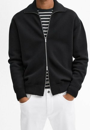 Homme portant une veste noire zippée par-dessus une chemise rayée noir et blanc avec un pantalon blanc, mains dans les poches.