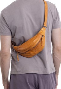 Mężczyzna noszący brązową skórzaną torbę na ramię, przylegającą do jego pleców, ubrany w jasnoszary t-shirt i ciemne szorty, stojący na białym tle.