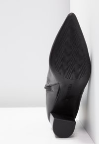 Botte en cuir noir à bout pointu avec semelle en caoutchouc texturée et petit talon carré, positionnée verticalement contre des murs d'angle blancs.