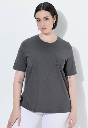 Frau mit kurzen dunklen Haaren, die ein dunkelgraues T-Shirt mit gemusterter Tasche und weiße Hose trägt, steht vor einer weißen Wand.
