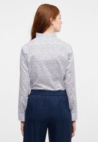 Weiße Bluse mit schwarzem Wirbelmuster, hochgeschlossenem Kragen und langen Ärmeln. Kombiniert mit dunkelblauen, elastischen Hosen mit Gummibund. Glatte Stofftextur.