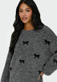 Grijze trui van pluizig materiaal met zwarte strikpatronen. Het ontwerp heeft een brede halslijn en losse mouwen.
