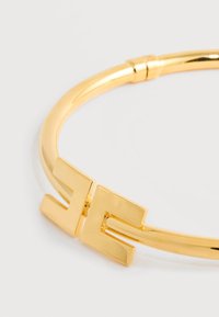 Βραχιόλι μπανγκλ σε χρυσή απόχρωση με λείο, στρογγυλεμένο σχέδιο, που διαθέτει γεωμετρικό κούμπωμα σε σχήμα "H" για ασφαλές κλείσιμο.