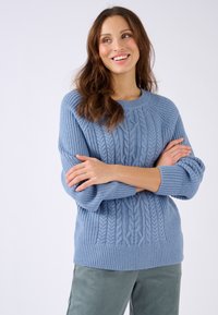 Lichtblauwe kabelgebreide trui met geribbelde manchetten en zoom, met een comfortabele pasvorm en zachte textuur, gecombineerd met groene ribfluwelen broek.