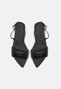 Mustat kärjelliset slingback-sandaalit, joissa on leveät pehmustetut remmit, joihin on kaiverrettu "HUGO"-logo, esitetty valkoista taustaa vasten.