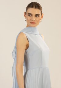 Robe sans manches bleu clair avec une jupe plissée, un col montant et un drapé fluide sur les épaules, fabriquée dans un tissu doux. Texture lisse et design minimaliste.