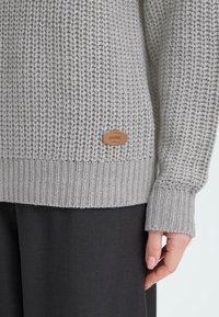 Grijze gebreide trui met een gestructureerd patroon, voorzien van een geribbelde onderkant en mouwen. Bruin label met de merknaam "OXMO" aan de onderkant.