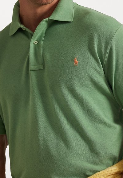 Άνδρας που φοράει ένα ανοιχτό πράσινο πόλο μπλουζάκι με ένα μικρό πορτοκαλί κεντητό λογότυπο στο στήθος, κρατώντας ένα κίτρινο ρούχο.