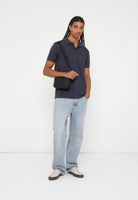 Marineblå polo skjorte, lyseblå løse jeans og cremefarvede sneakers. Model bærer en lille sort crossbody taske med lynlåslukning.