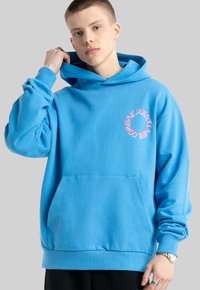 Niebieska bluza z kapturem, wykonana z mieszanki bawełny, z kieszonką z przodu, ściągaczami przy mankietach oraz różowym nadrukowanym okrągłym logo po lewej stronie klatki piersiowej.