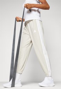 Beige Sporthose mit weißen seitlichen Streifen, kombiniert mit weißen Sportschuhen. Die Hose hat einen elastischen Bund und eine lockere Passform.