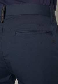 Gros plan de la poche arrière d'un pantalon bleu marine avec une poche fendue horizontale et une passants de ceinture, porté avec un haut sombre.