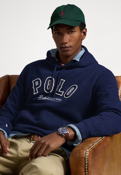 Vyras dėvintis žalią kepuraitę, tamsiai mėlyną Polo Ralph Lauren džemperį su gaubtu ant džinsinio marškinių, rudas kelnes ir laikrodį, sėdintis ant rudos odinės fotelio.