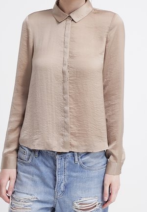 Femme portant un chemisier en satin beige à manches longues boutonné, associé à un jean en denim bleu clair déchiré.