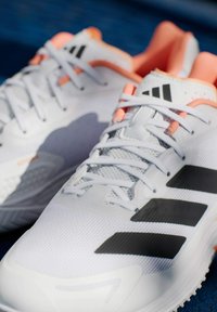 Λευκά αθλητικά παπούτσια με μαύρες ρίγες, ελαφρύ γκρι πλέγμα άνω μέρος, πορτοκαλί λεπτομέρειες, υφασμένη σόλα και κλείσιμο με κορδόνια.